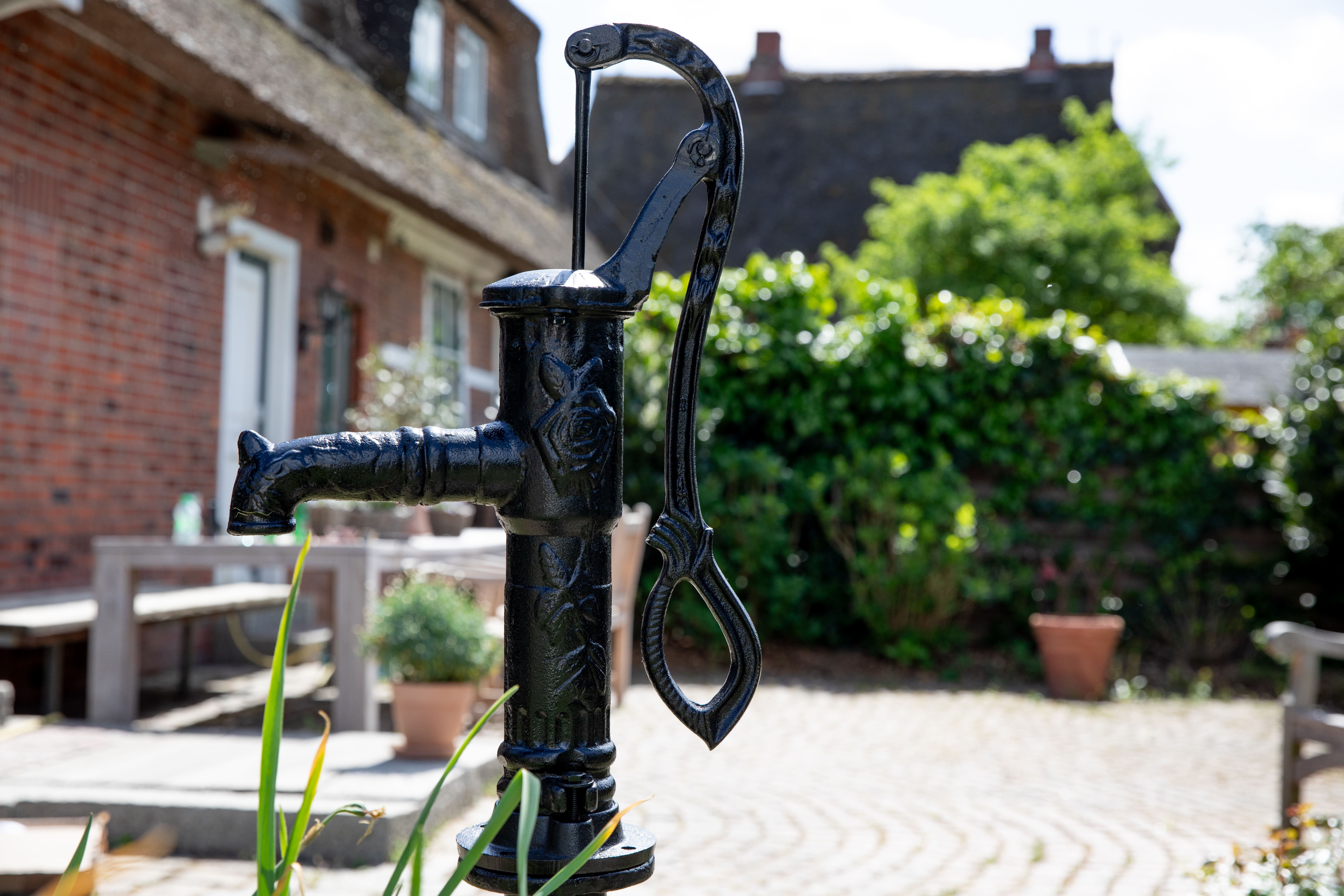 Schwarze Schwengelpumpe aus Gusseisen vor Backsteinhaus im Garten mit Kopfsteinpflaster